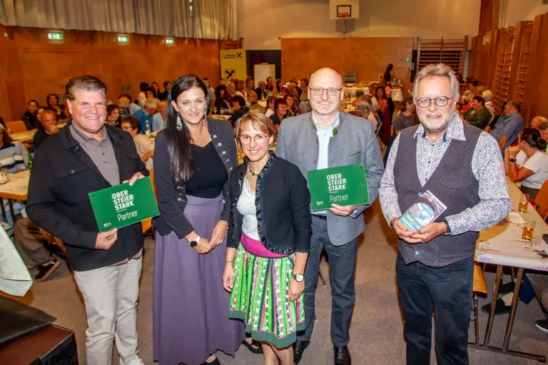 Kraubath setzte ein Highlight zum Booktober Zwei Frauen und drei Männer mit Schildern und einem Buch in Händen stehen in einem Turnsaal, im Hintergrund voll besetzte Tische