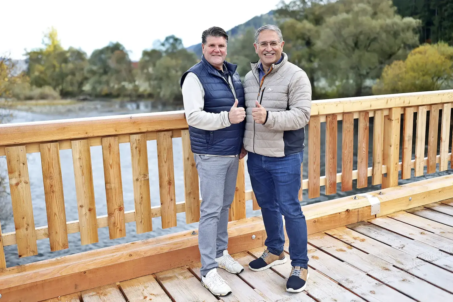 Zwei Männer stehen mit erhobenen Daumen auf einer Holzbrücke