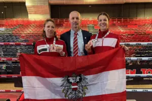 Zwei Frauen in rot-weißen Trainingsjacken halten ihre Bronzemedaillen in die Kamera. Zwischen ihnen steht ein Mann im dunklen Anzug mit rot-weiß gestreifter Krawatte. Gemeinsam halten sie eine österreichische Flagge vor einem Boxring. Im Hintergrund sind rote und schwarze Zuschauertribünen zu sehen.