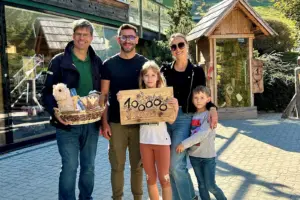 Drei Erwachsene und zwei Kinder stehen mit einer Holztafel und einem Geschenkskorb auf einem Platz vor mehreren kleinen Holzgebäuden