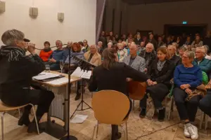 Zwei Musikerinnen sitzen vor einem zahlreich erschienenen Publikum. Eine Frau singt in ein Mikrofon, die andere spielt ein Streichinstrument.