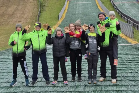 Erfolgreicher Sommersaison-Abschluss der Schispringer Eine Gruppe von Kindern und einem Erwachsenen posiert auf dem Auslauf einer Skisprungschanze für ein Gruppenfoto. Mehrere Kinder halten kleine Holzpokale in die Höhe. Die Personen tragen Winterjacken und Hauben, einige davon leuchtend grüne Vereinsjacken. Im Hintergrund sind die Skisprungschanzen zu sehen.