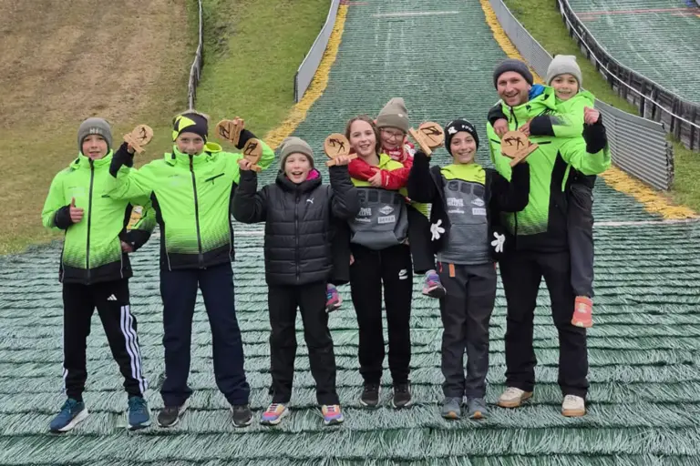 Erfolgreicher Sommersaison-Abschluss der Schispringer Eine Gruppe von Kindern und einem Erwachsenen posiert auf dem Auslauf einer Skisprungschanze für ein Gruppenfoto. Mehrere Kinder halten kleine Holzpokale in die Höhe. Die Personen tragen Winterjacken und Hauben, einige davon leuchtend grüne Vereinsjacken. Im Hintergrund sind die Skisprungschanzen zu sehen.