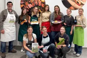 Neun junge Frauen und ein Mann mit Schürzen präsentieren Lebensmittel und ein Buch
