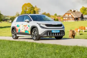 Ein hellgraues Auto der Kategorie Kompakt-SUV mit bunt beklebten Türen fährt auf einer Landstraße von links nach rechts