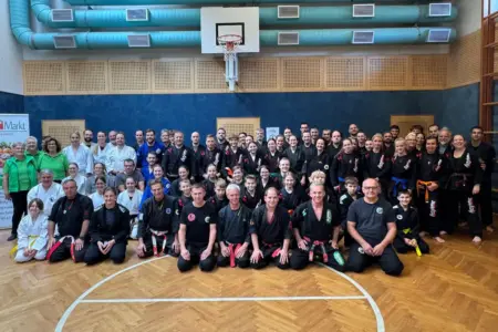 Kampfsportler sammelten für guten Zweck Große Gruppe von Frauen, Männern und Kindern in unterschiedlichen Kampfsportanzügen, überwiegend in Schwarz und Weiß, posiert in mehreren Reihen für ein Gruppenfoto in einer Turnhalle. Einige Personen knien oder sitzen vorne, andere stehen dahinter. Im Hintergrund sind eine blaue Wand und ein Basketballkorb zu sehen.