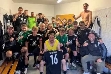 Großer Jubel in der Kabine: Die Fußballer des SV Stanz und ihr Betreuerteam feiern ausgelassen ihren Erfolg. Im Mittelpunkt präsentiert ein Spieler stolz ein Trikot mit der Nummer 16.