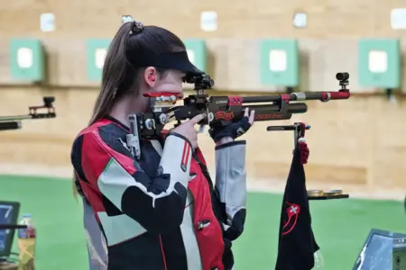 Eine Sportschützin mit Pferdeschwanz, schwarzem Visier und rot-schwarz-weißer Schießjacke zielt mit einem Luftgewehr durch ein Zielfernrohr. Sie hält das Gewehr mit beiden Händen. Im Hintergrund ist eine Schießhalle zu sehen.