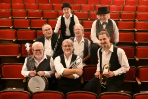 Sieben Männer in weißen Hemden und schwarzen Gilets mit Musikinstrumenten auf roten Theatersesseln.