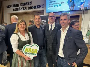 Eine Frau mit einem grünen Herz in der Hand zeigt sich mit drei Männern in dunkeln Sakkos.